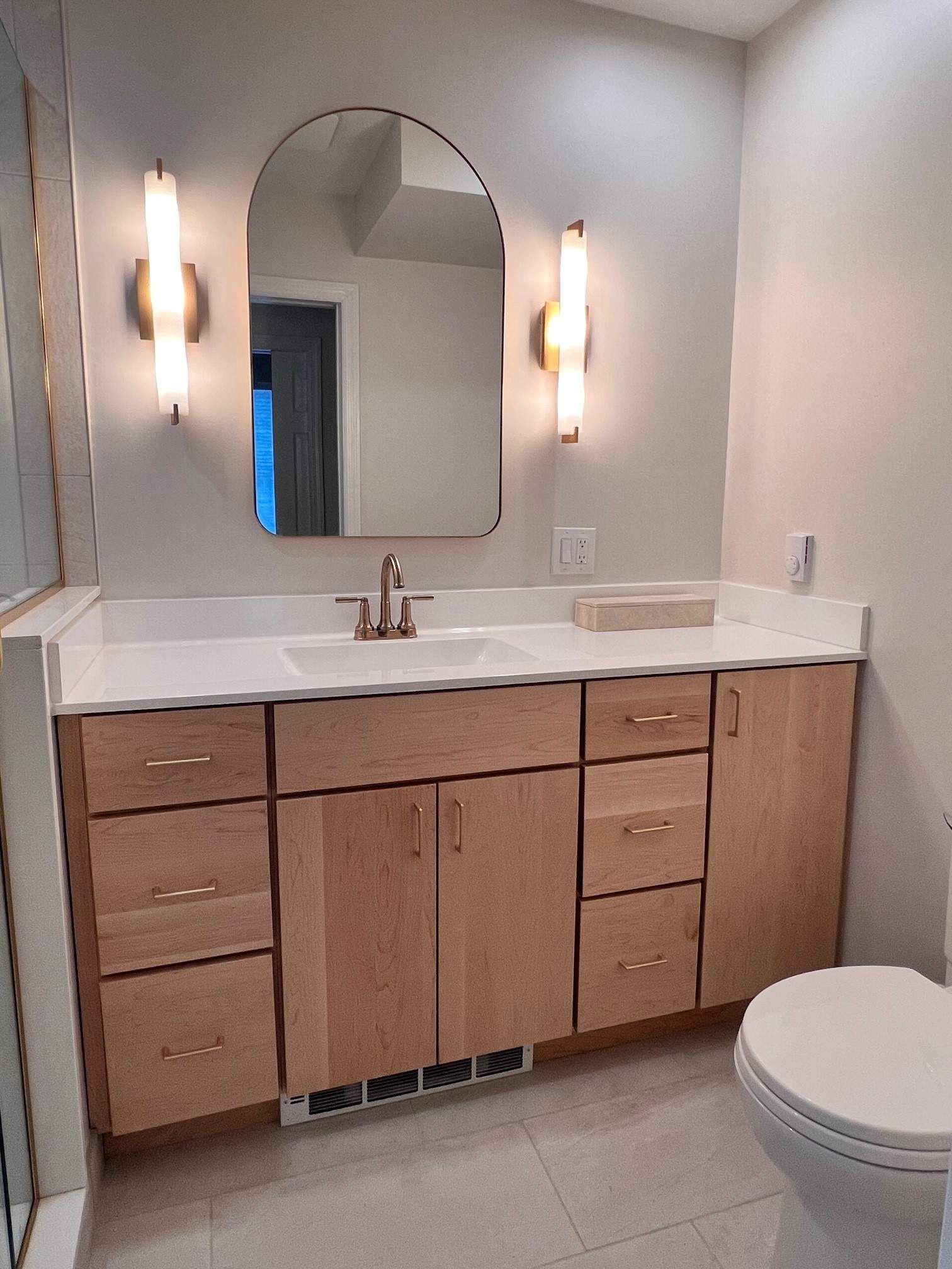 Bathroom vanity with light wood cabinets, white countertop, arched mirror, and sconces.