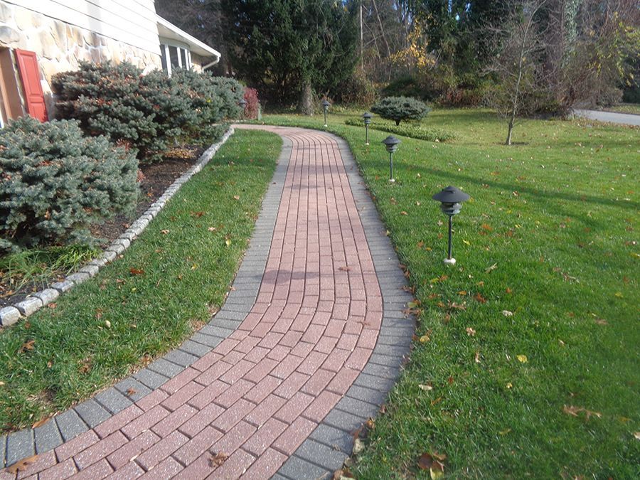 Brick pathway winds through green grass, bordered by shrubs and garden lights.