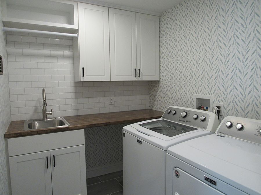 Laundry room with white cabinets, appliances, and sink, and patterned wallpaper.