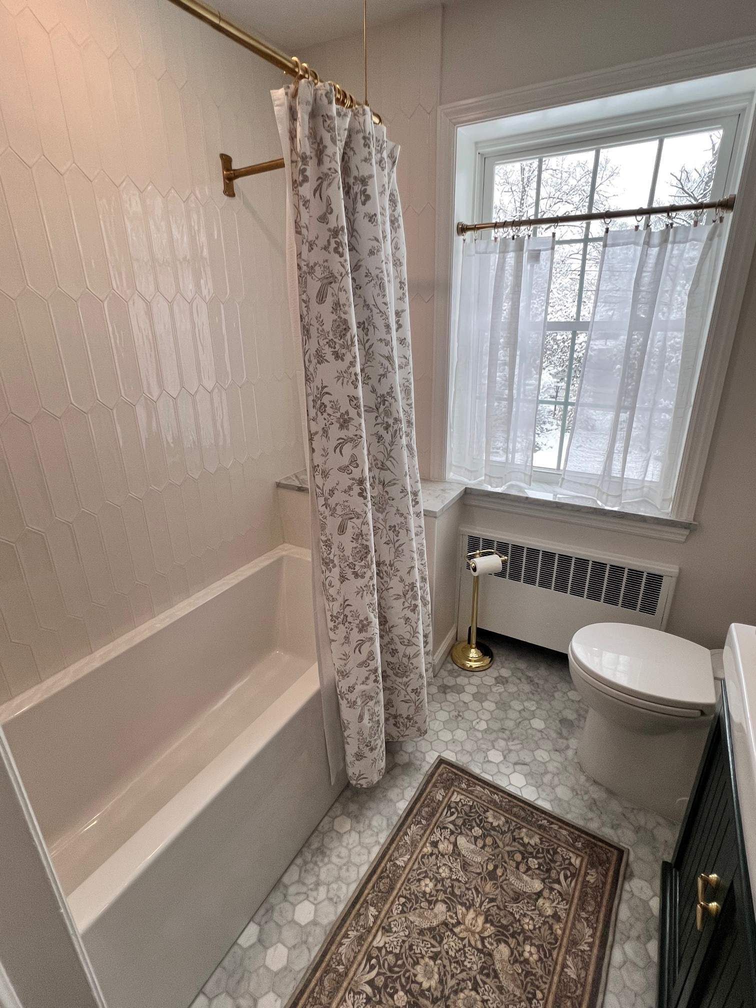 Bathroom with a white bathtub, patterned shower curtain, and a window with a sheer curtain. Gray and white hexagonal floor tiles.