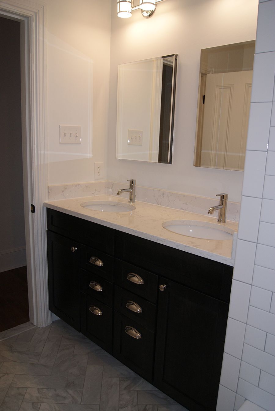 Bathroom with black vanity, white countertop, two sinks, mirrors, and white tile.