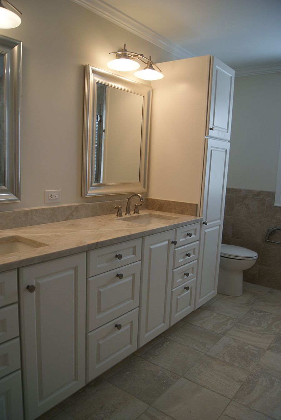 Bathroom with white cabinets, double sink vanity, tall cabinet, and a toilet. Light stone-like tile floor.