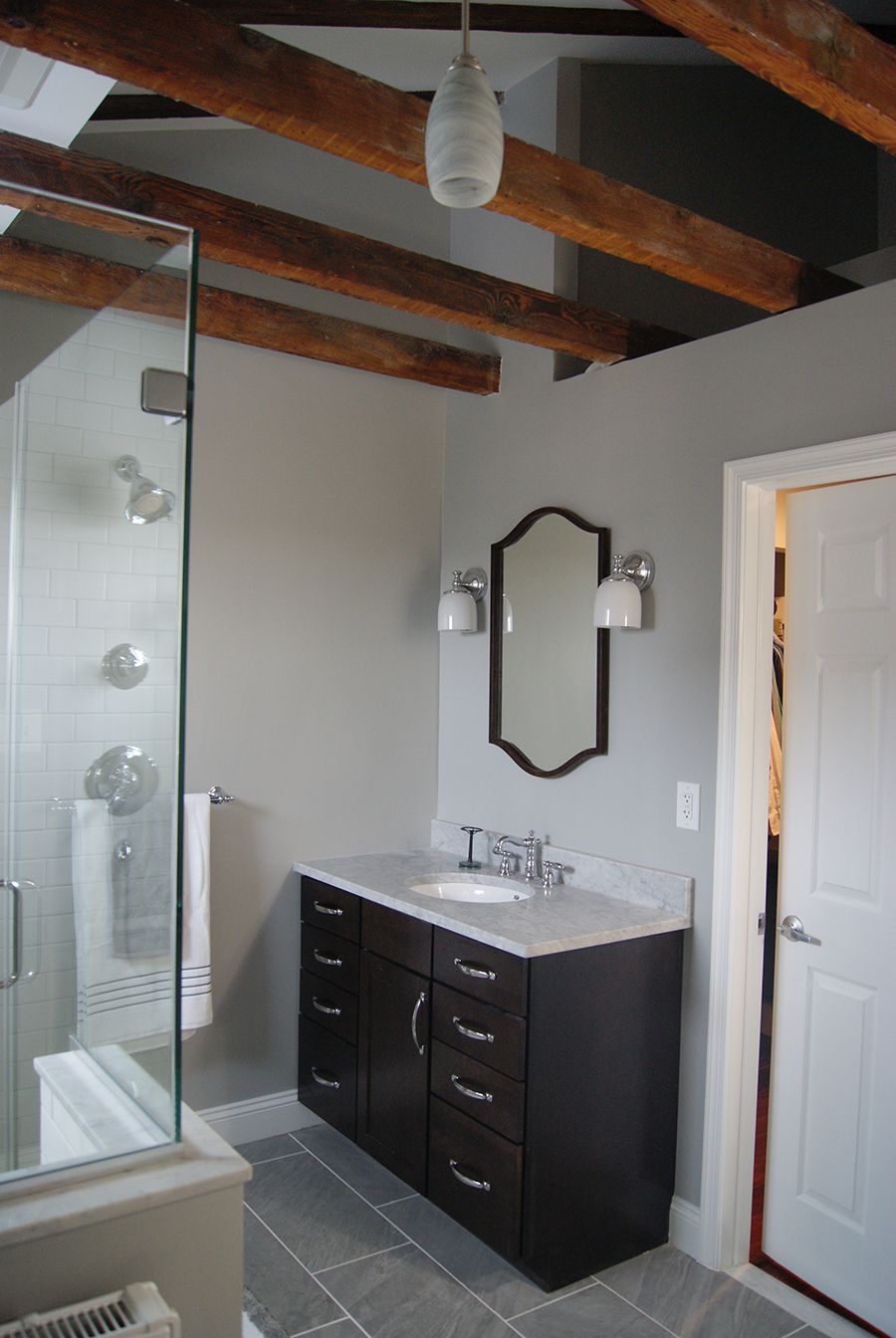Bathroom with a glass shower, dark vanity, gray walls, exposed wooden beams, and a white door.