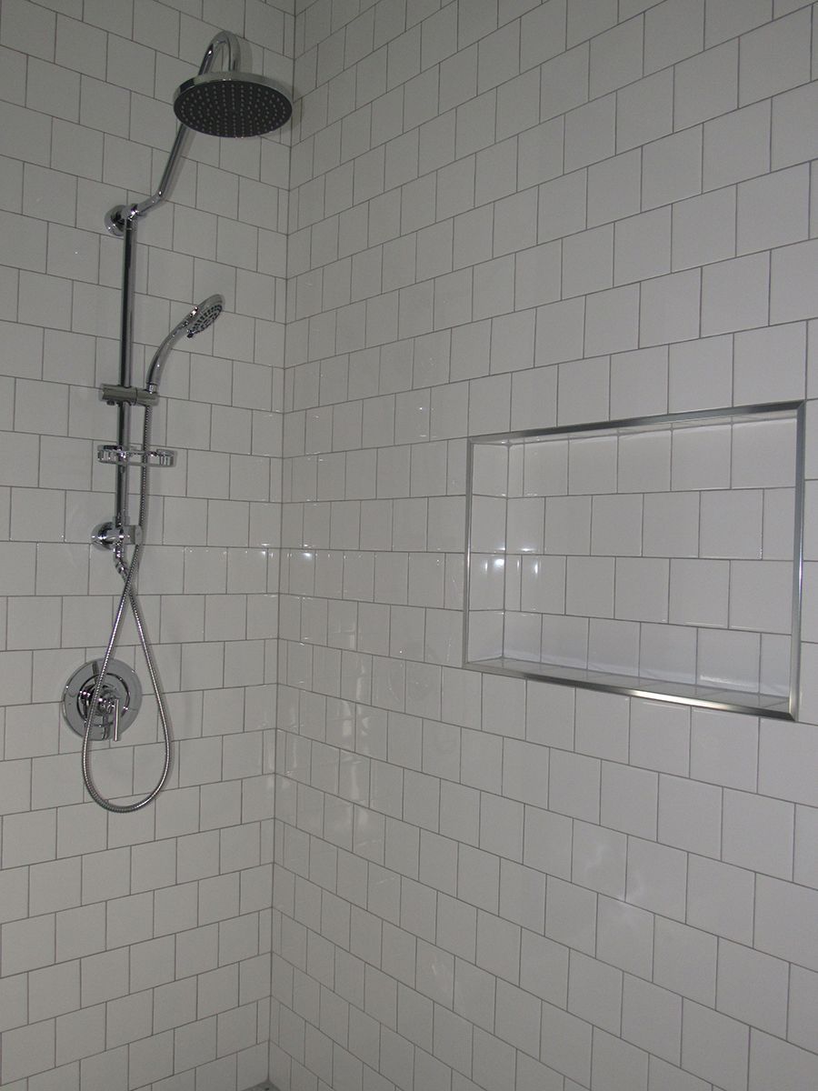 White-tiled shower with shower head and wand; a recessed shelf is built into the wall.