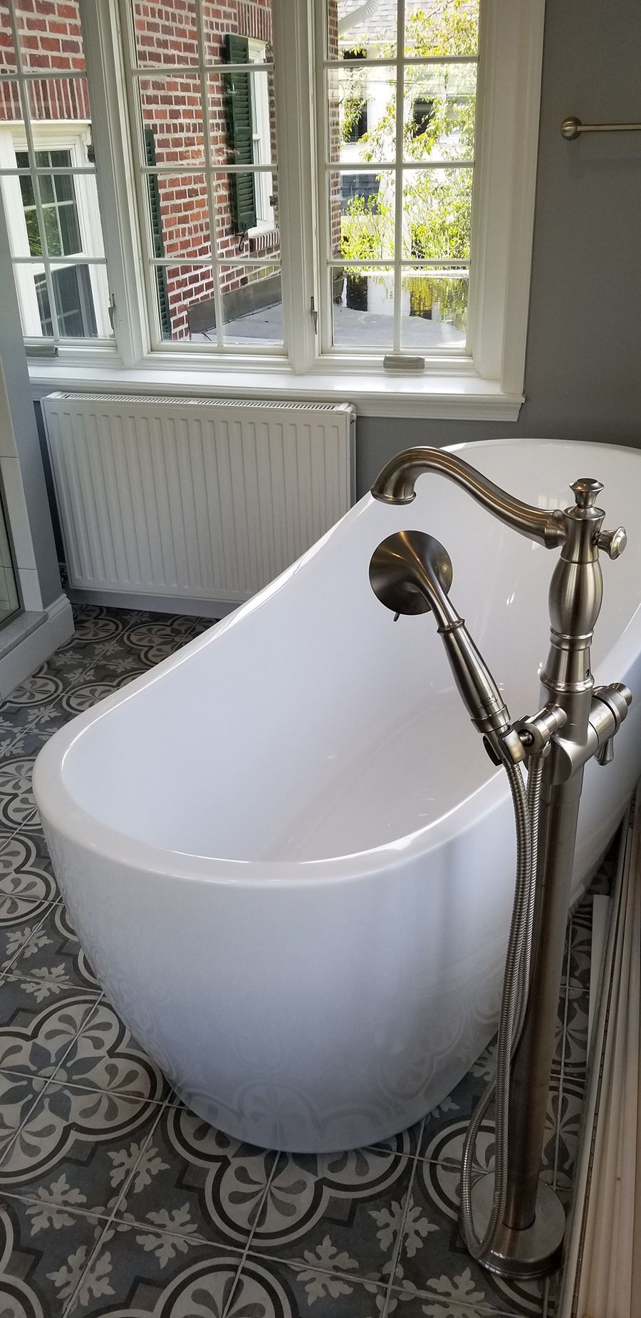 White freestanding bathtub in a bathroom with patterned floor, silver faucet, and window with a view of a brick building.