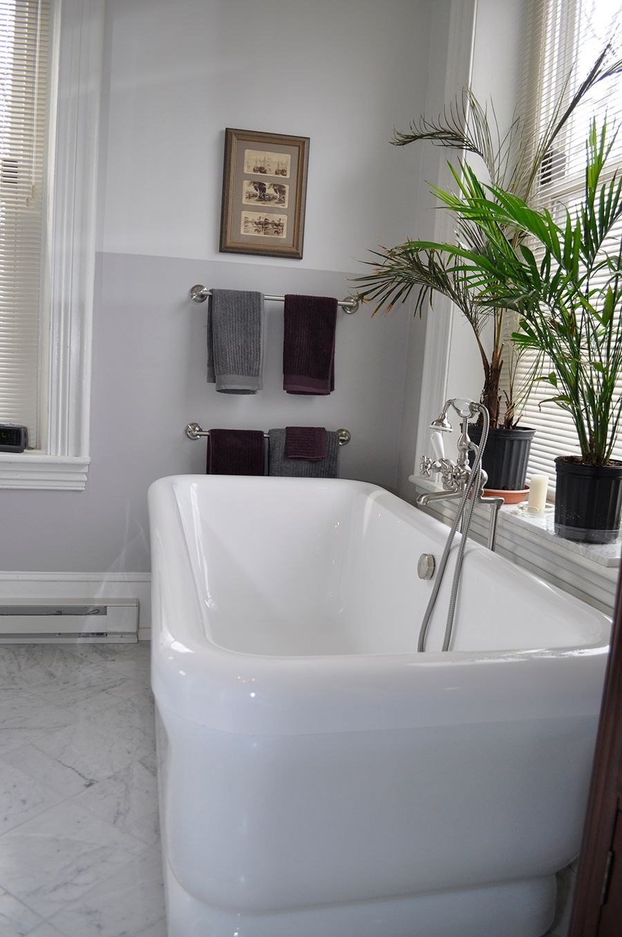 Bright white bathtub in a light grey bathroom with towels, artwork, and potted plants near a window.