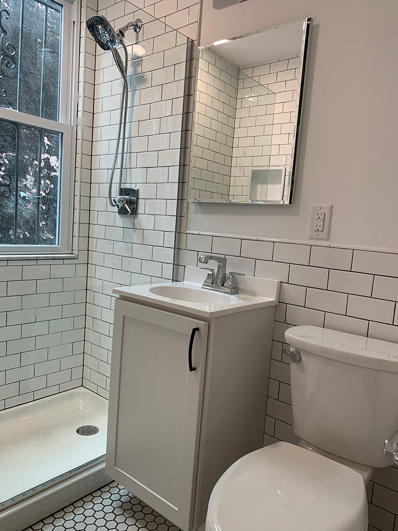 Small, modern bathroom with white tile and fixtures; sink, toilet, shower, and mirror visible.