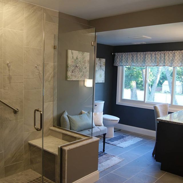 Bathroom with a tiled shower, toilet, window, and small bench. Gray walls and flooring.