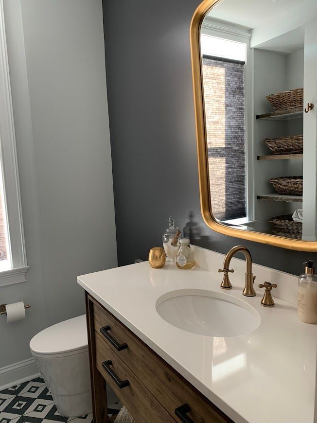 Bathroom with white countertop, gold mirror, wooden vanity, gray and white walls, and black and white patterned floor tiles.