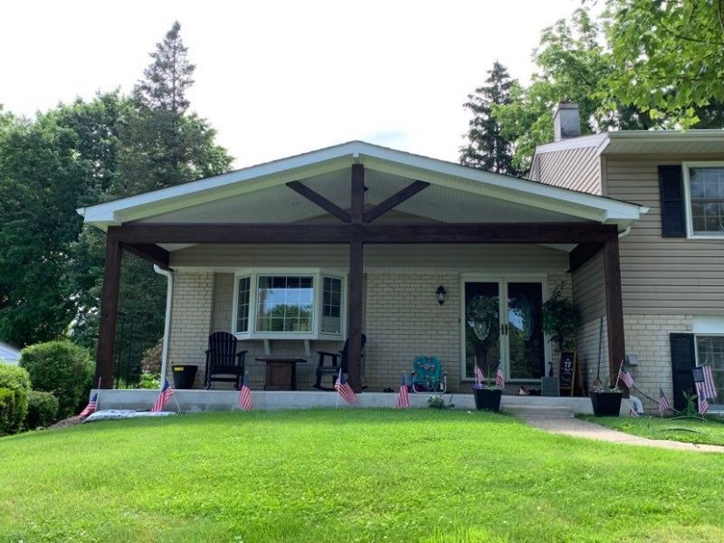 A house with a porch. Brown beams frame the porch roof, supported by columns. American flags decorate the lawn.