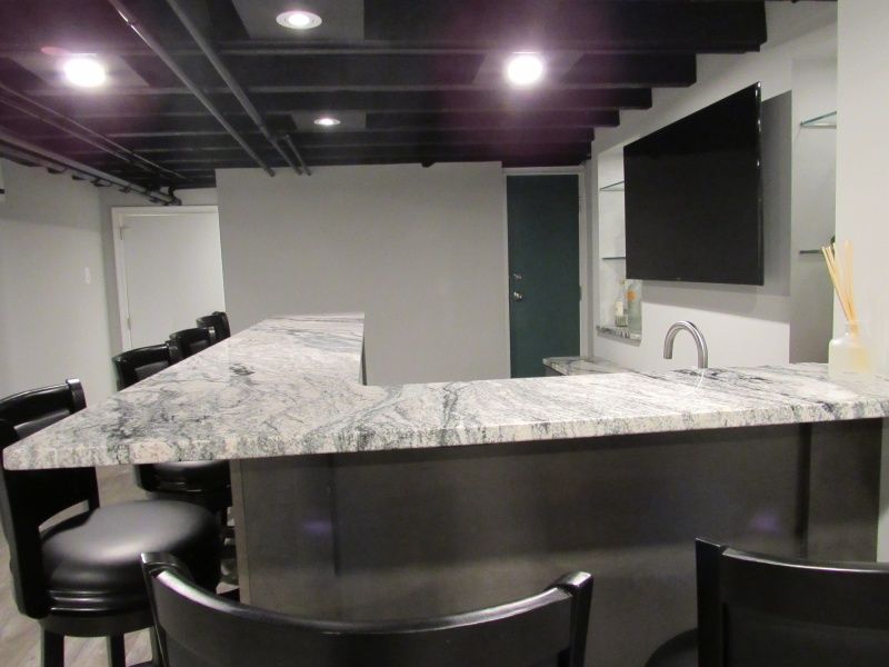 A granite-topped bar with black stools and TV in a basement with dark ceiling.