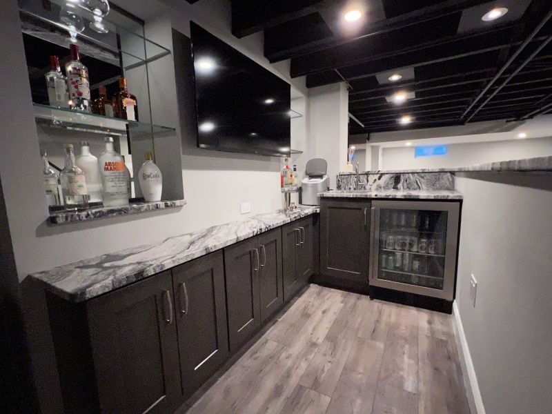 Basement bar with gray cabinets, marble countertops, a mini-fridge, a TV, and liquor bottles on shelves.