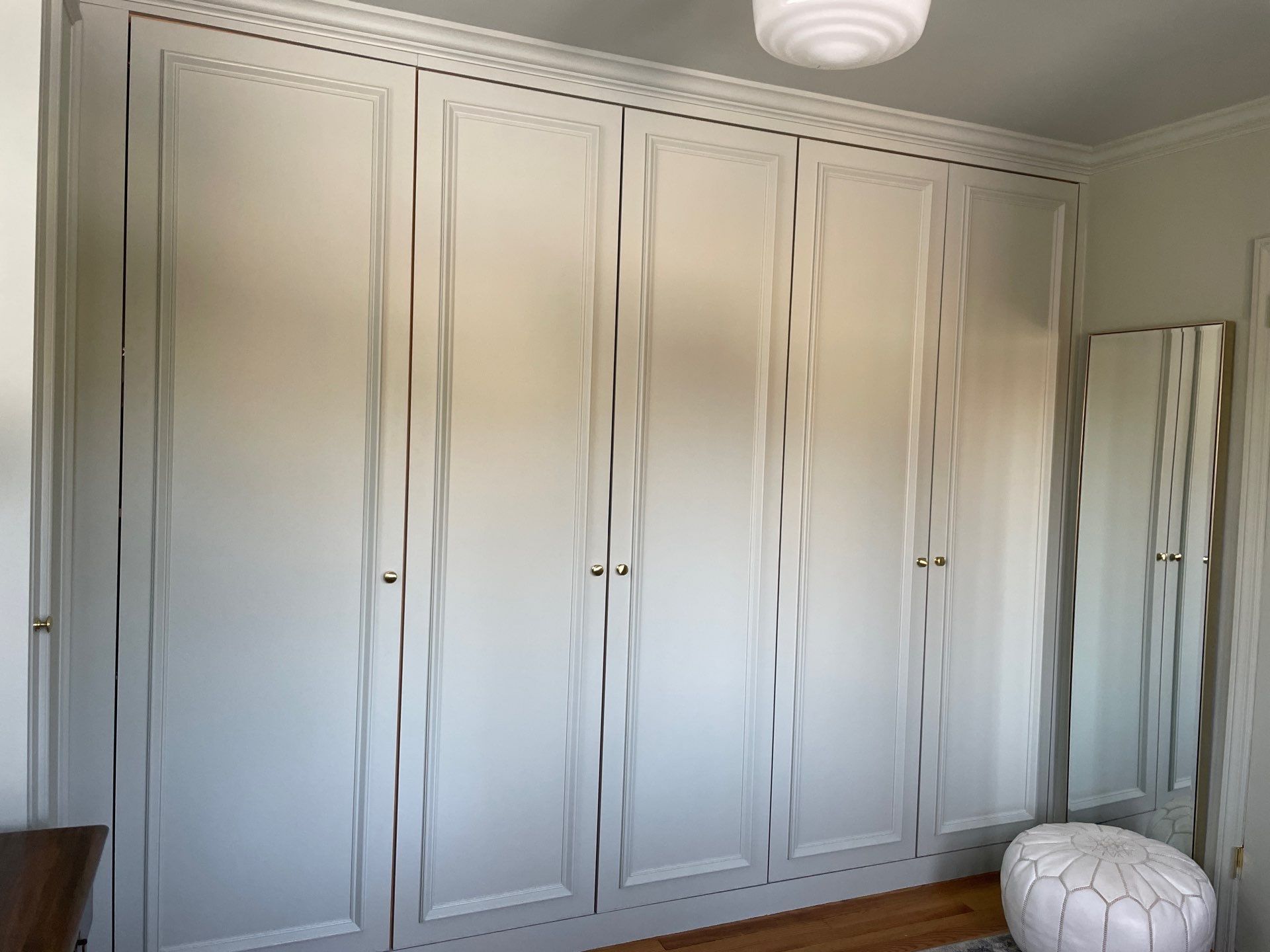 Built-in white cabinets with decorative molding and wavy trim. A mirror and ottoman are next to the cabinet.