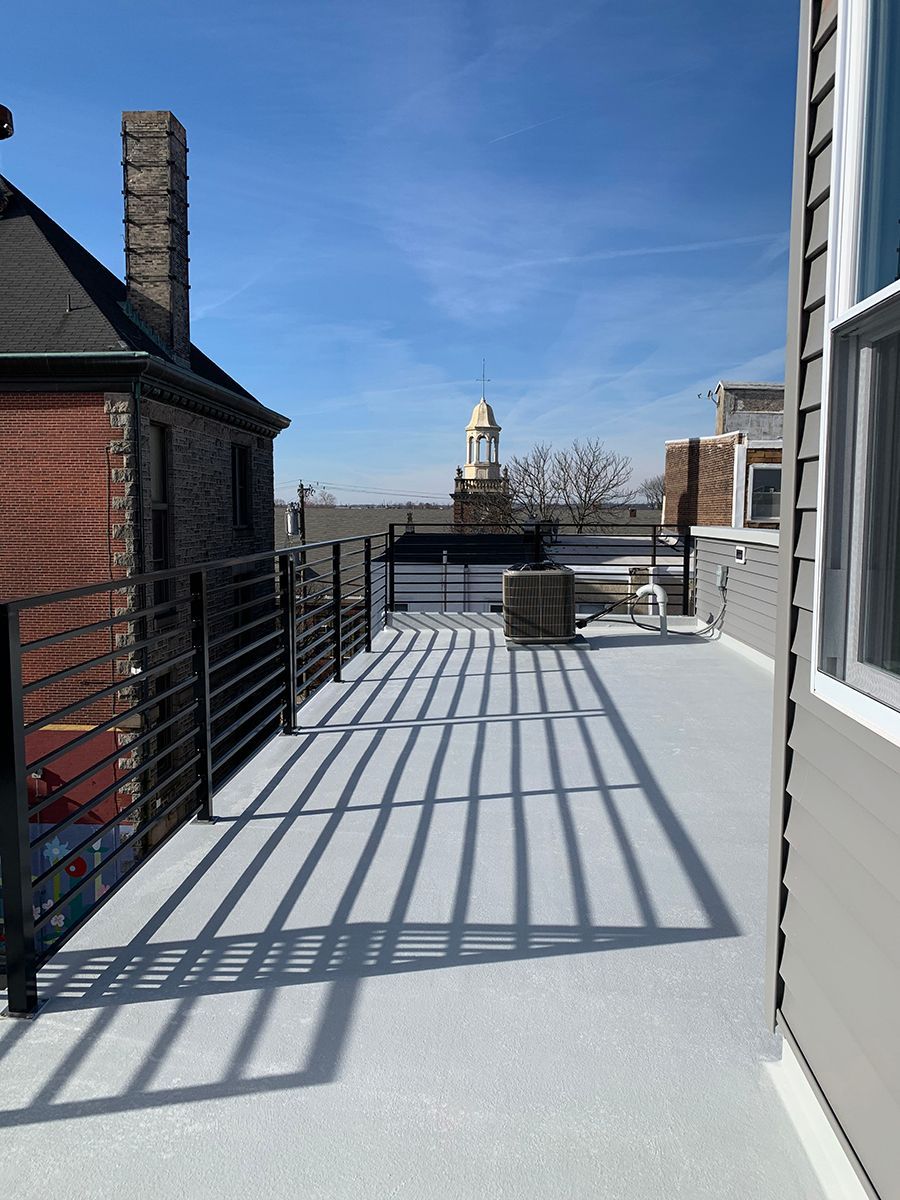 Rooftop with metal railing, view of buildings, blue sky. Grey surface casts shadows from the railing.