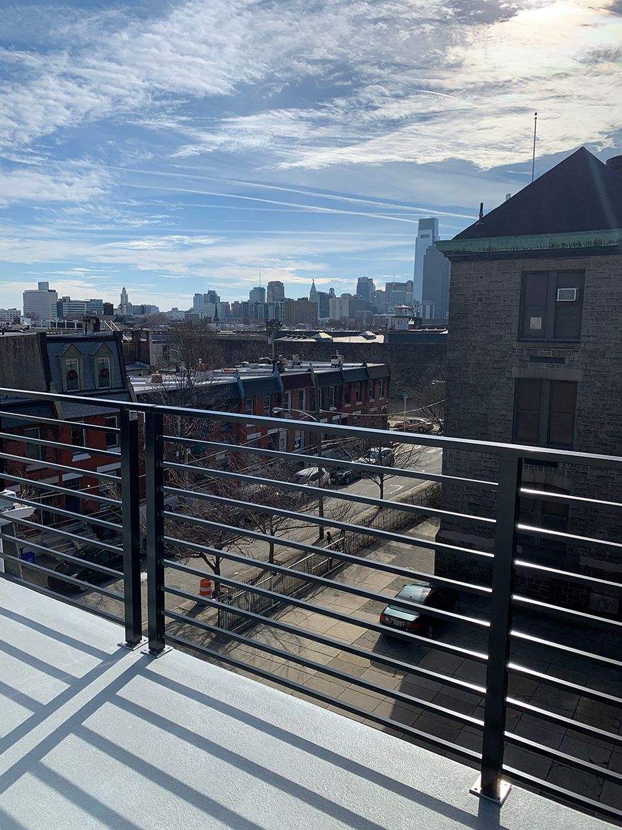 Cityscape view from a rooftop, featuring a skyline, street, and building with a metal railing. Blue sky with clouds.