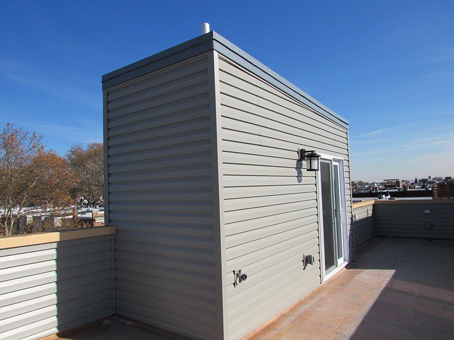 Gray-sided rooftop structure with a door, light fixture, and spigots on a concrete deck against a blue sky.
