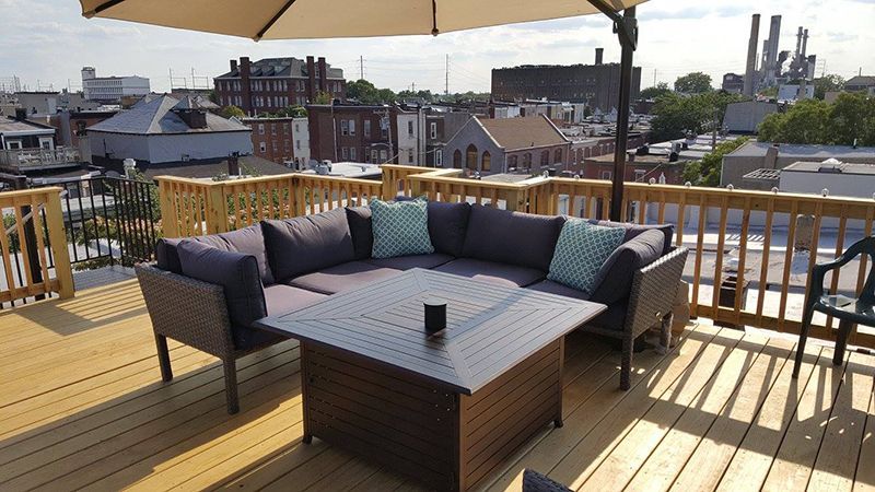Rooftop deck with seating area: dark sectional sofa, square fire table, tan umbrella, cityscape background.