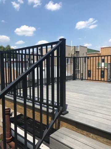 Black railing and deck on a rooftop, sunny day. Buildings in the background.