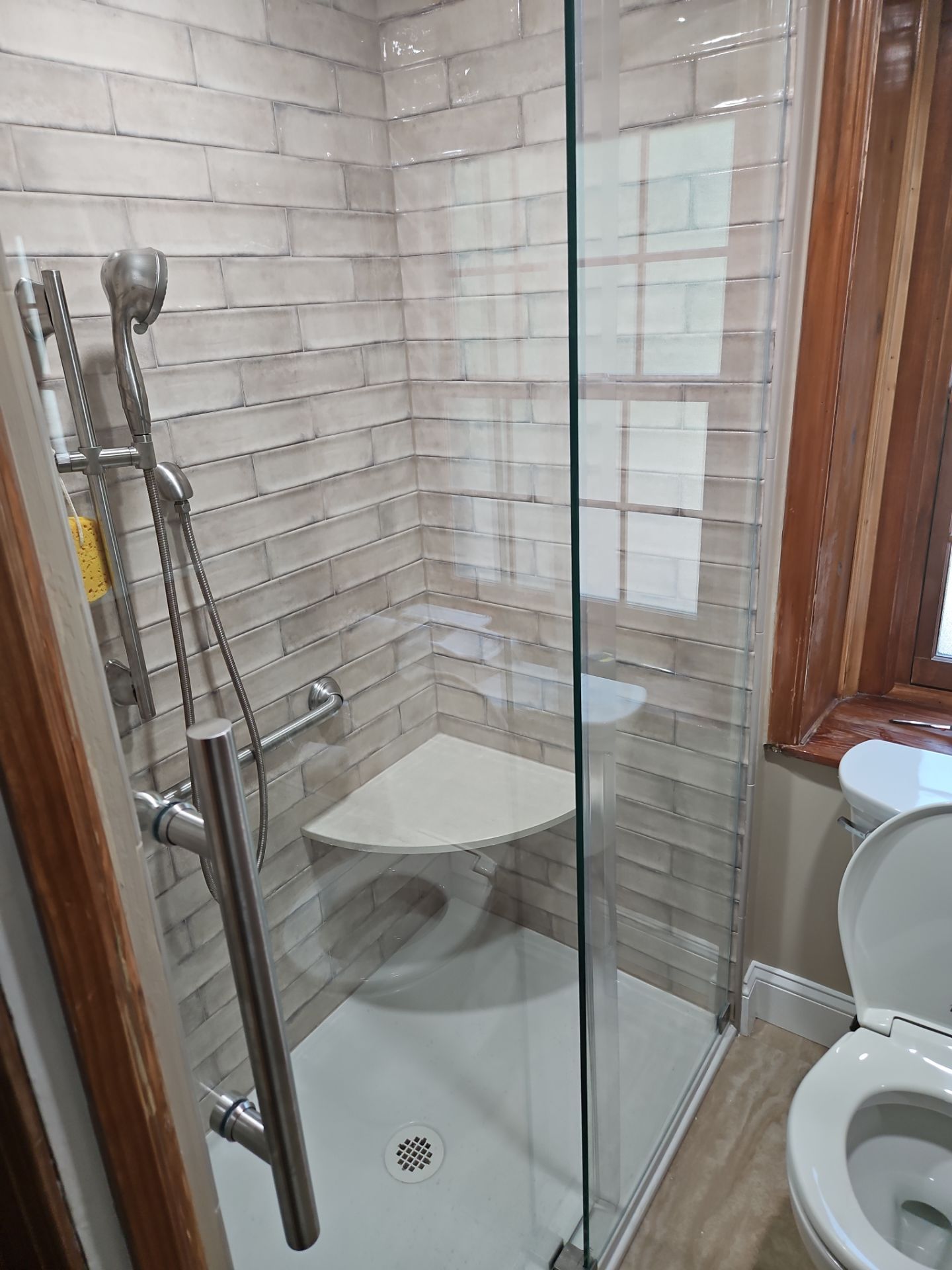 Accessible shower with corner seat, grab bar, and glass door. Tile walls, toilet visible.