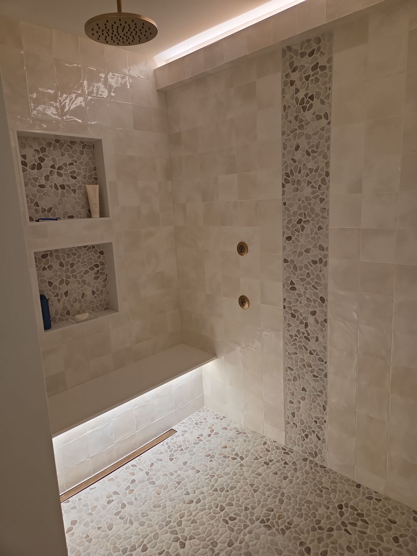 Modern tiled shower with built-in bench and recessed shelves. Features stone mosaic accents, a rain showerhead, and gold fixtures.