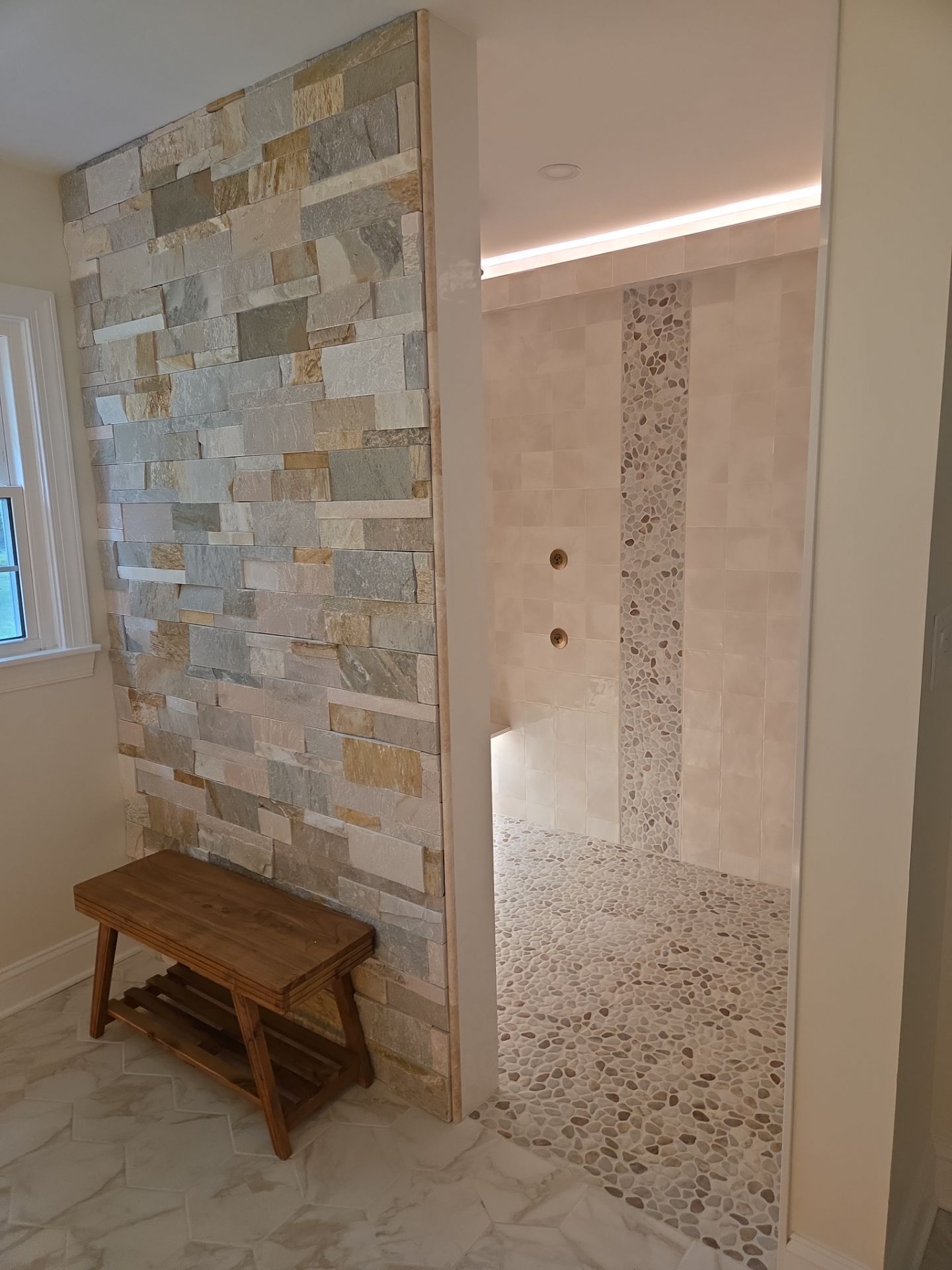 A modern bathroom with a stone accent wall, wooden bench, and walk-in shower.