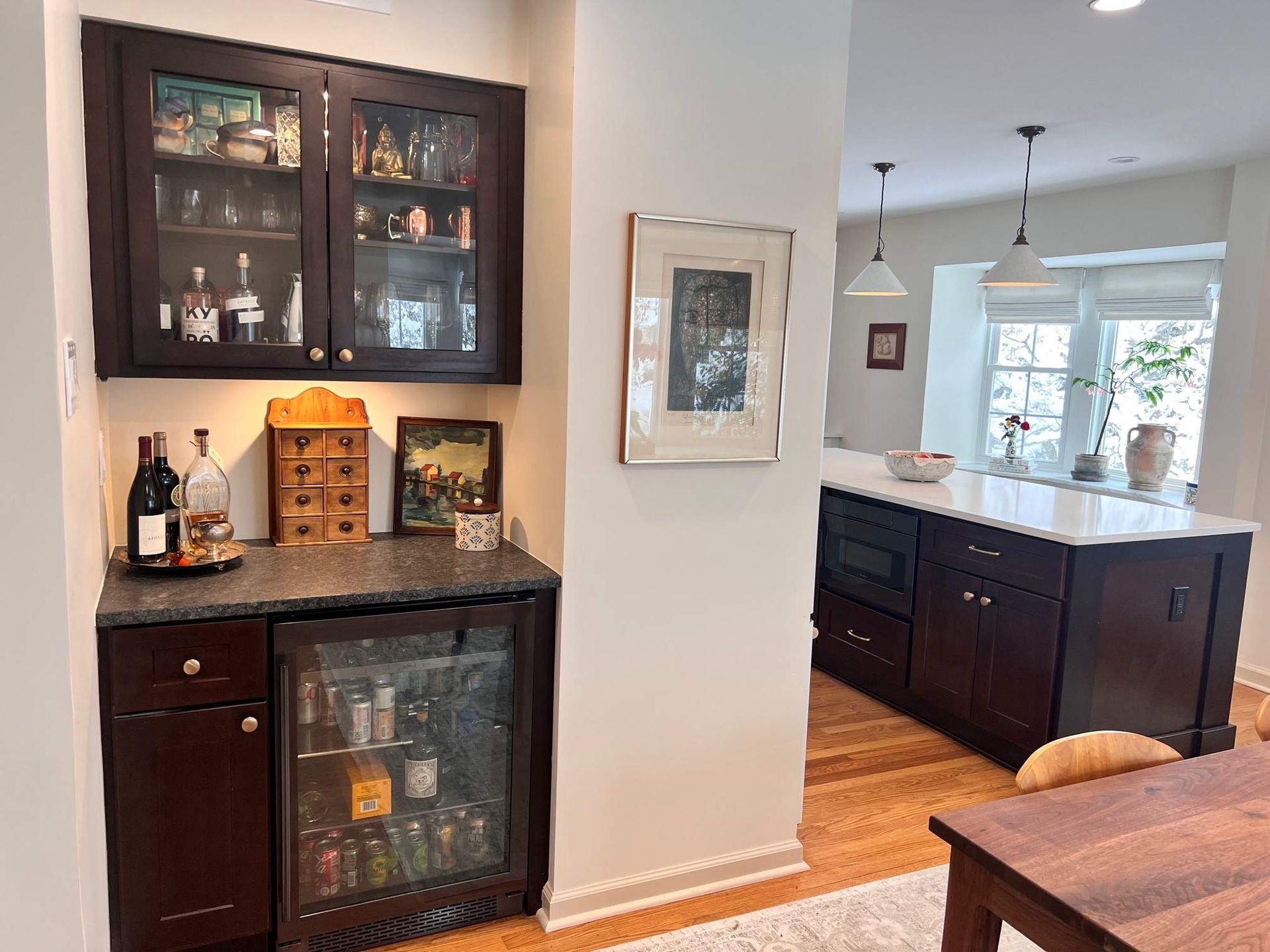 Dark wood bar area with cabinet, mini-fridge, and liquor bottles next to a kitchen island.