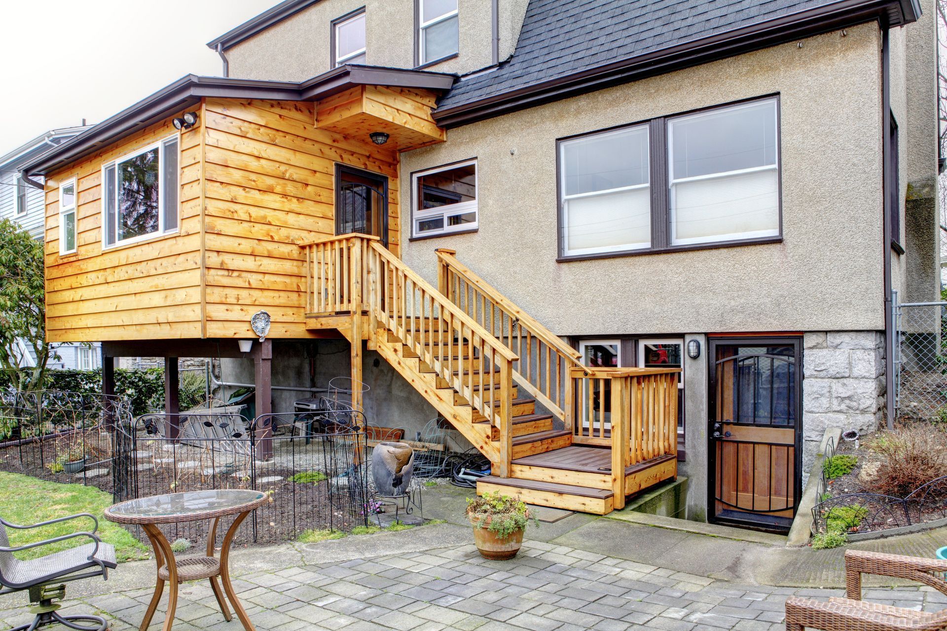 Back of a house with wooden deck, stairs, and patio. Light brown wood siding on a raised addition.