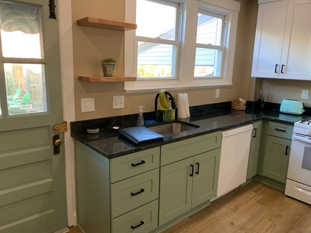 Kitchen with sage green cabinets, black countertops, a white dishwasher, and light-colored wooden shelves.