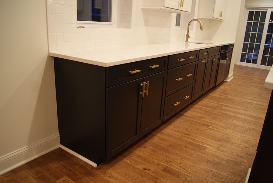 Black kitchen cabinets with gold hardware and white countertop on wood floor.
