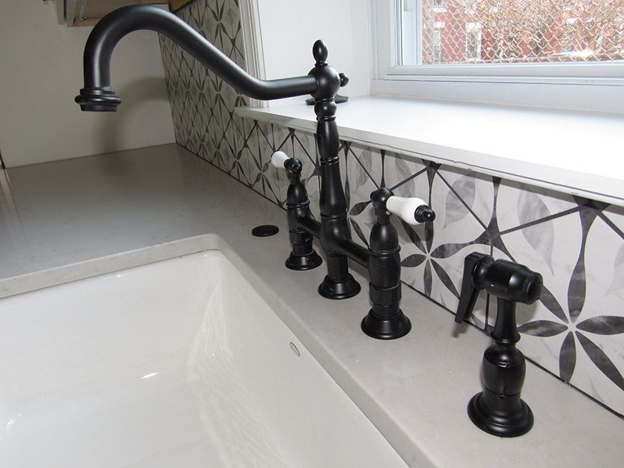 Black faucet and sprayer on a white countertop by a patterned backsplash and window.