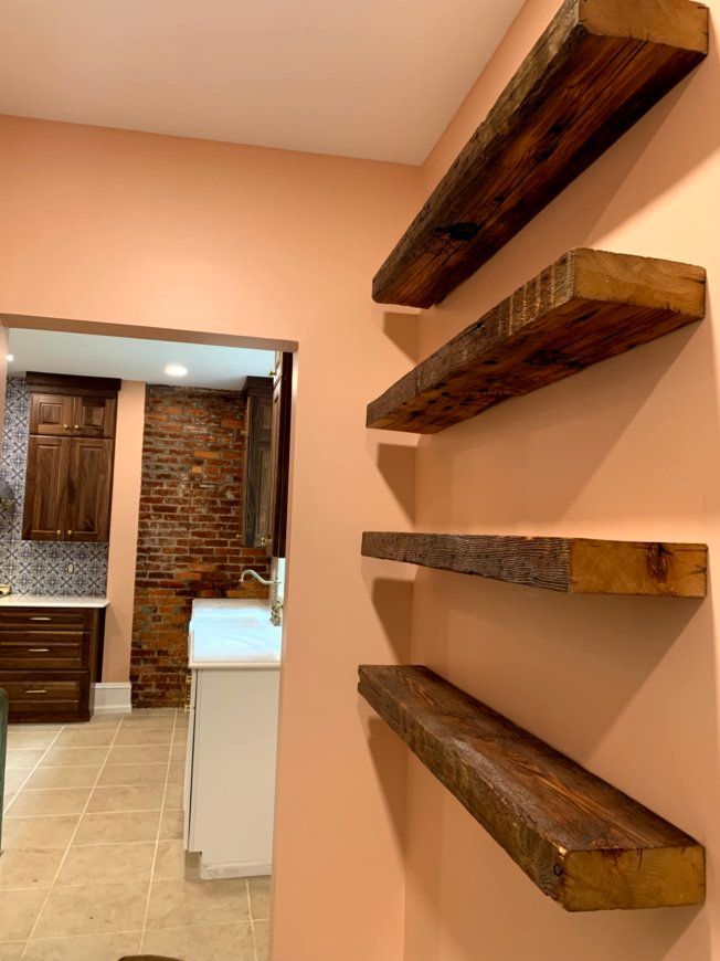 Four rustic wooden shelves on a peach-colored wall; kitchen with brick wall and cabinets visible in the background.