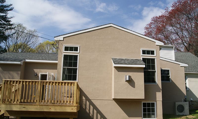 Rear view of a two-story beige house with a wooden deck and several windows under a blue sky.