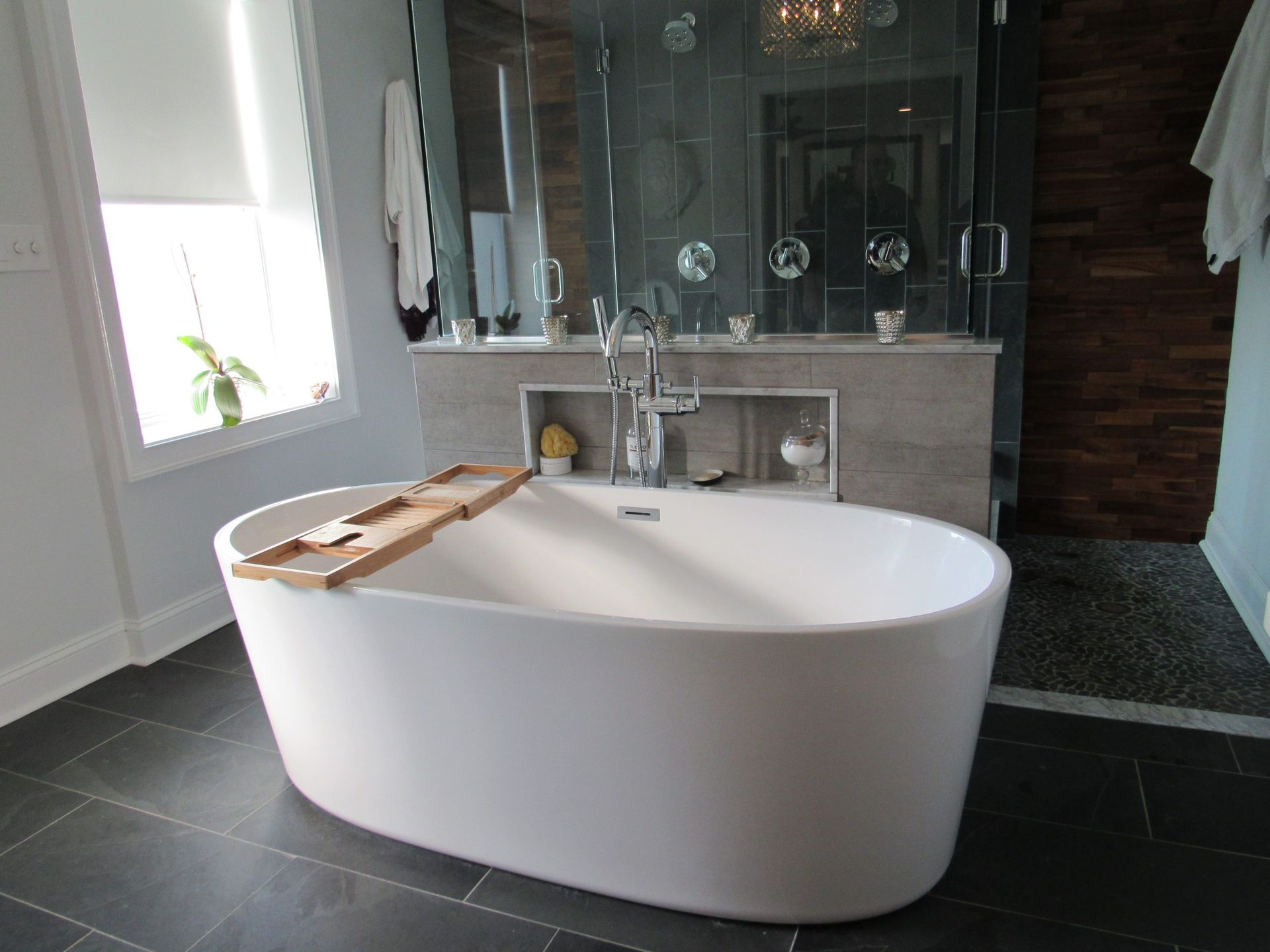 Modern white oval bathtub in a bathroom, with a wooden tray, dark tile floor, and a glass shower.