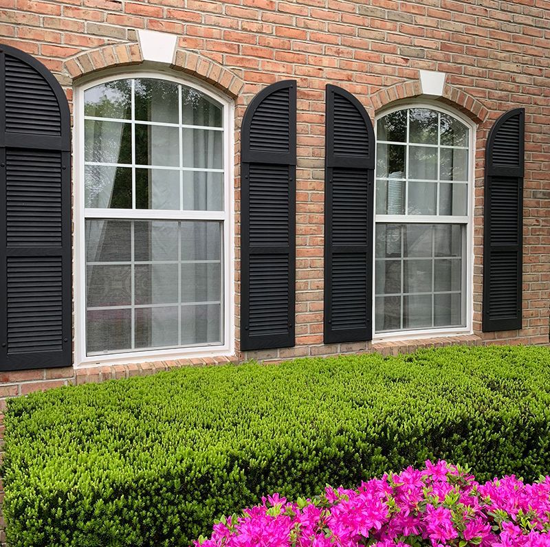 Brick building with arched windows and black shutters. Green bush in front with pink flowers.