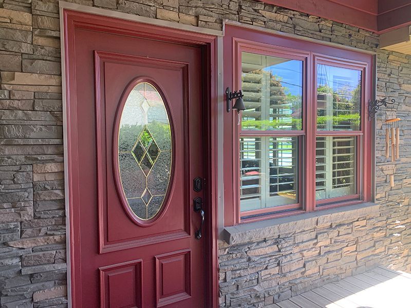 Red front door with oval glass and adjacent window with shutters, set in a stone facade.