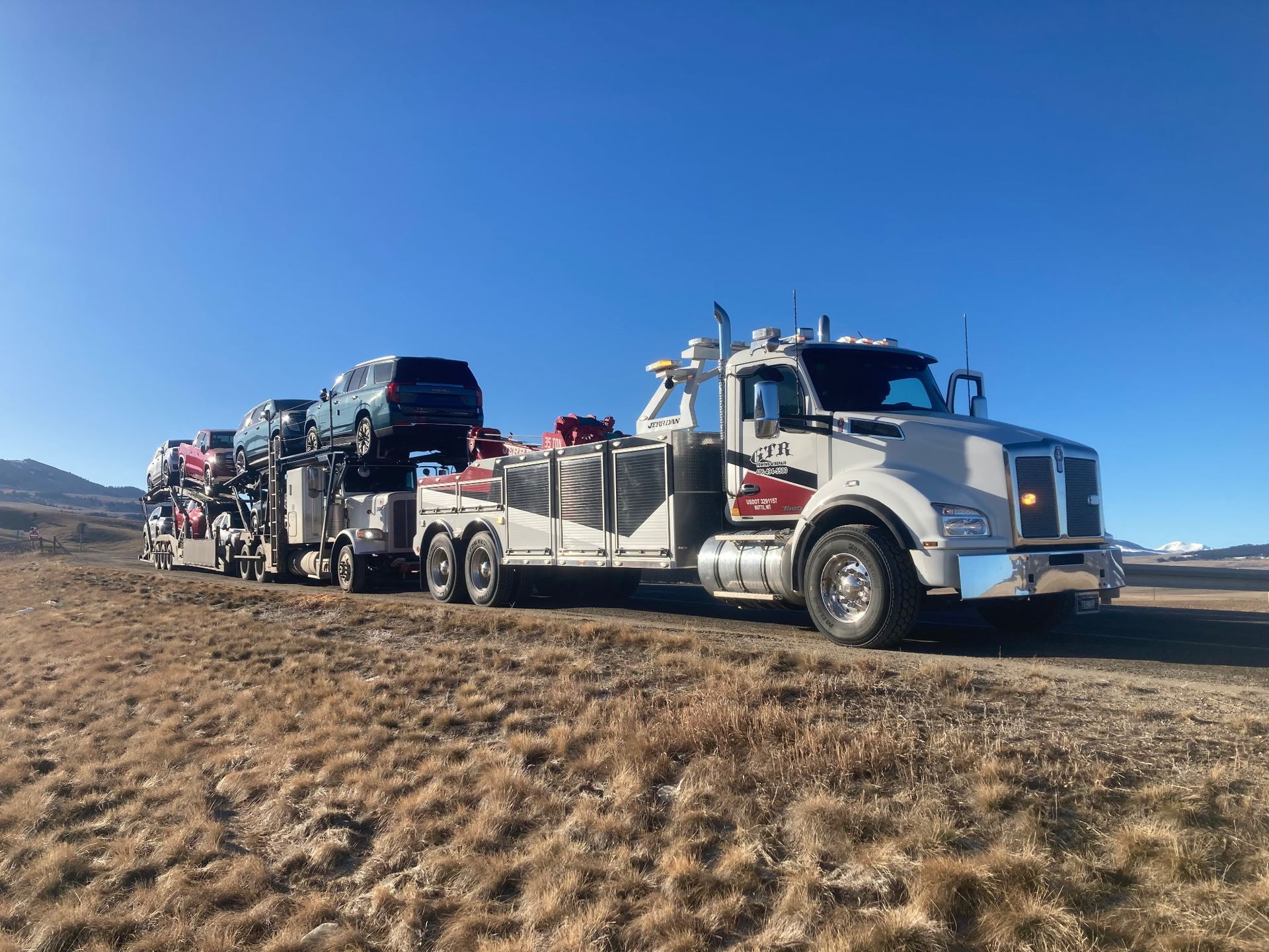 A white semi-truck hauling several cars on a flatbed trailer across a dry, grassy field under a clear blue sky.