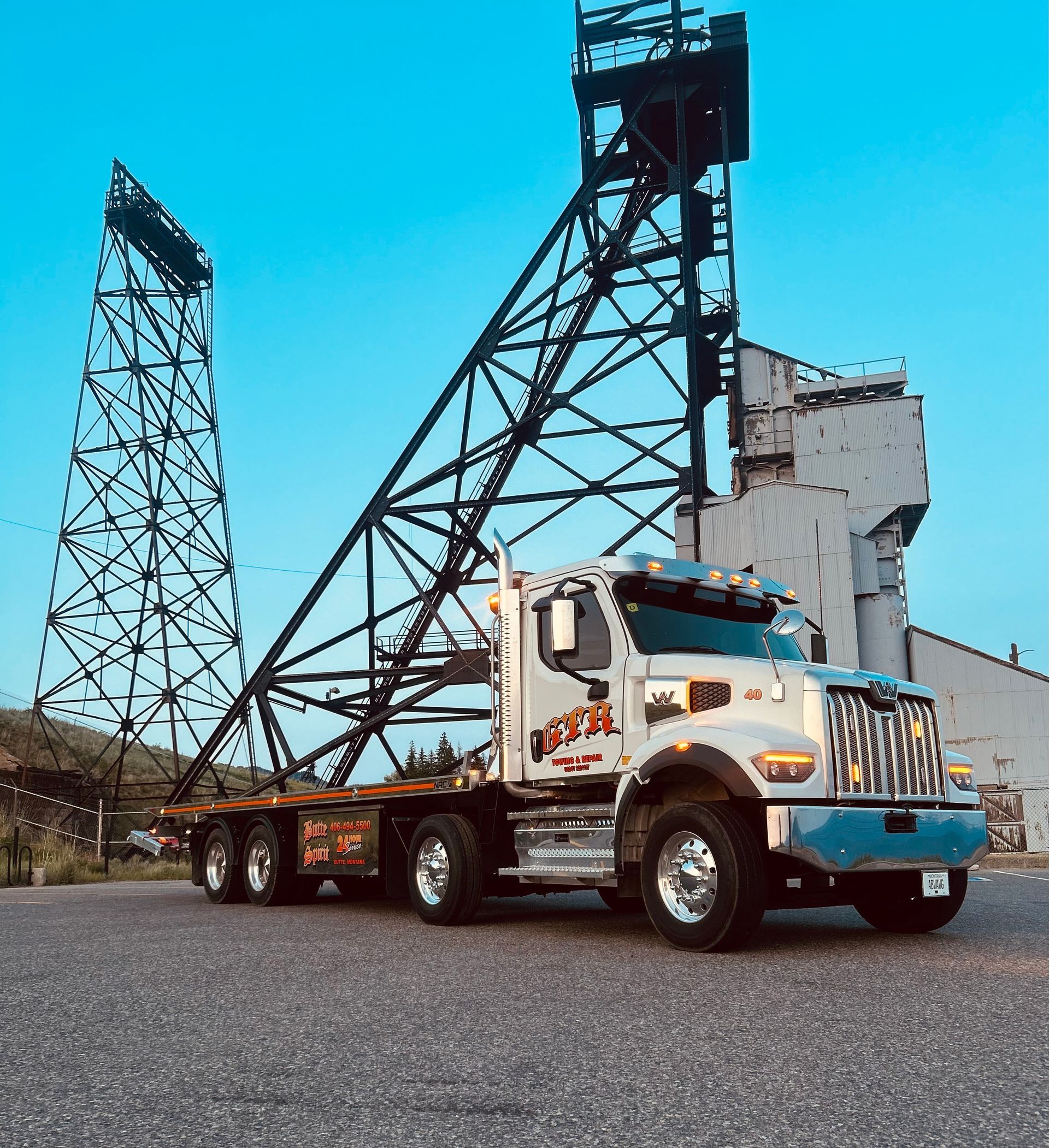 A white flatbed truck parked on a gravel lot in front of a tall, dark steel mining headframe structure under a blue sky.