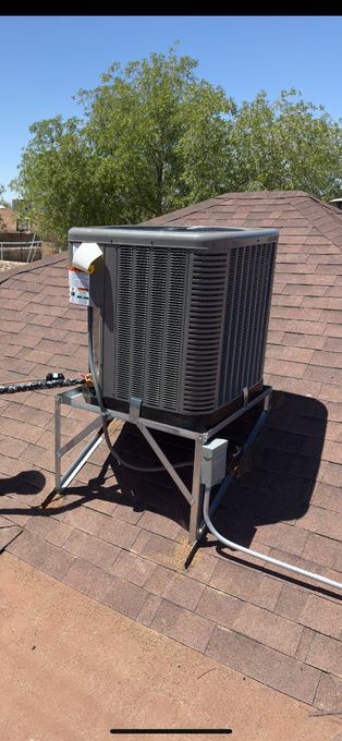 An air conditioning unit sits on a metal stand on a rooftop, blue sky and green trees in the background.