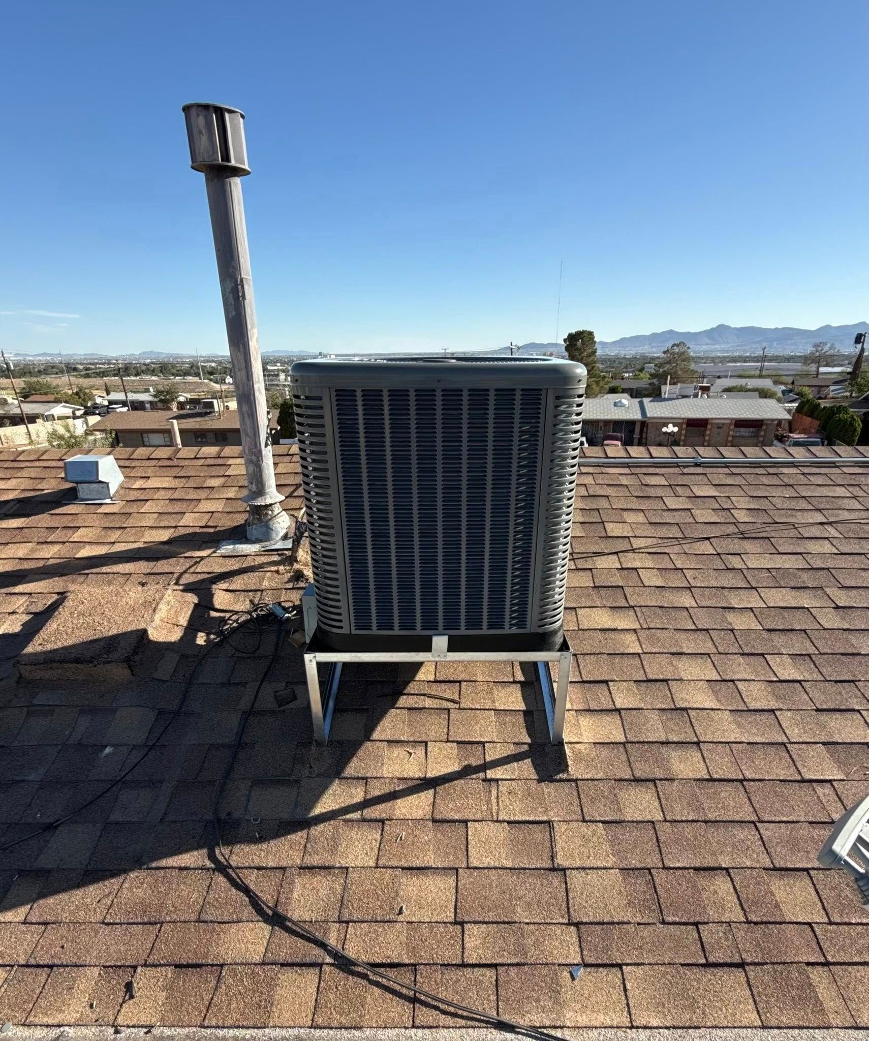 HVAC unit on a brown shingle roof with a chimney, clear blue sky and distant buildings.