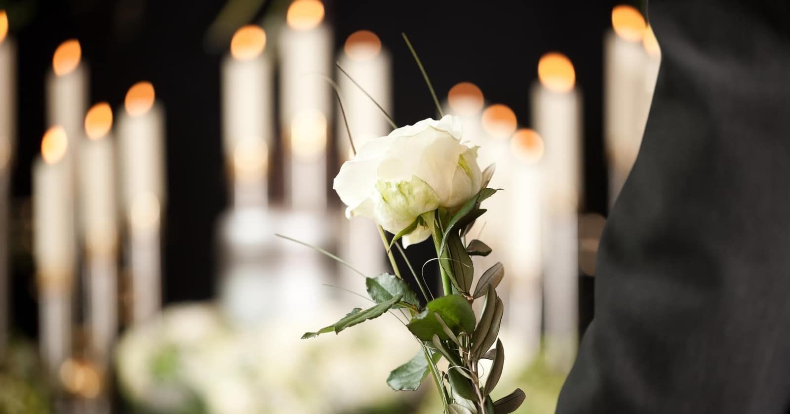 A person is holding a white rose in front of candles at a funeral.