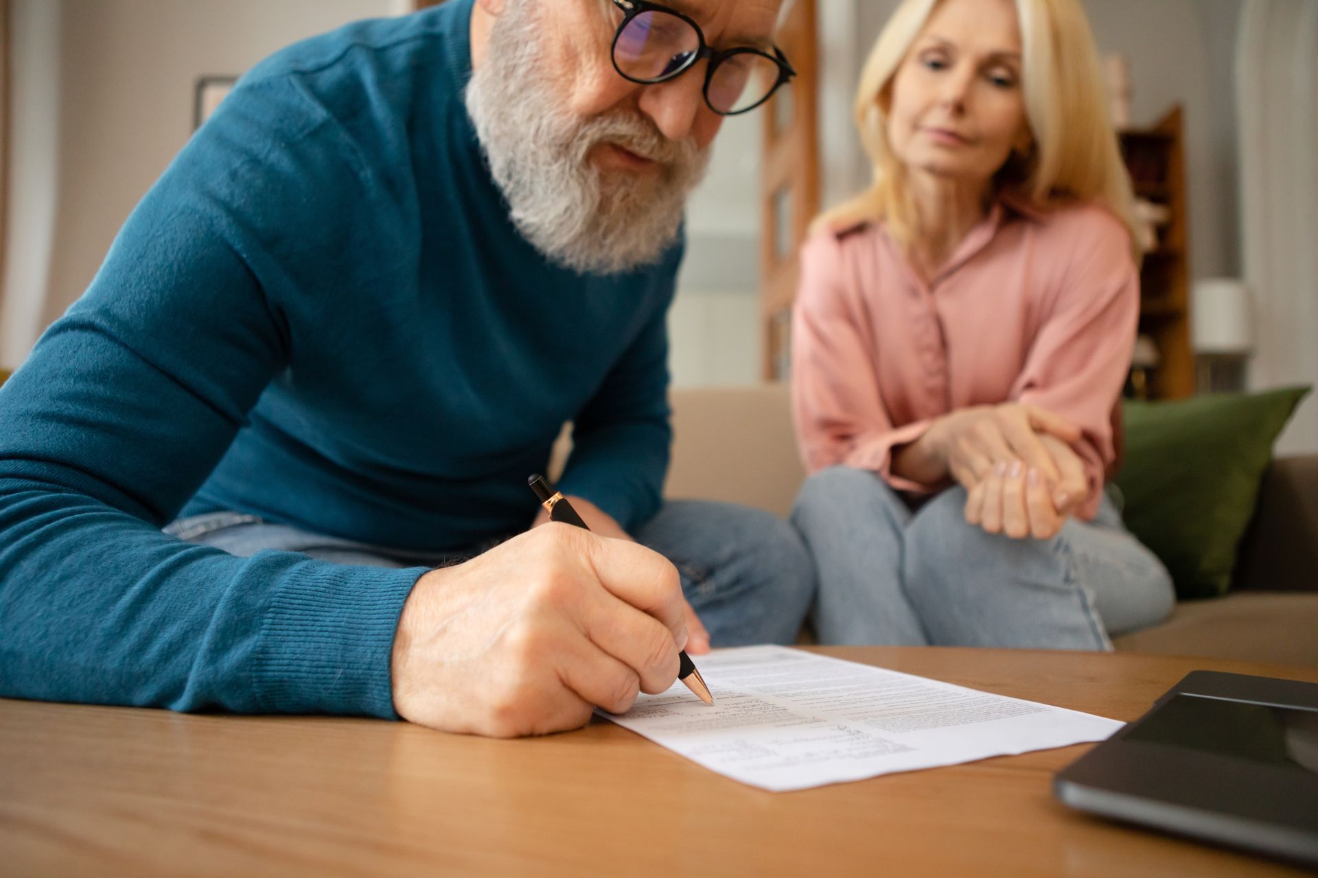 A man is signing a document while a woman sits on a couch.