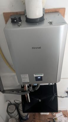 Rinnai tankless water heater mounted on a wall. Silver casing, white exhaust pipe, and digital display.
