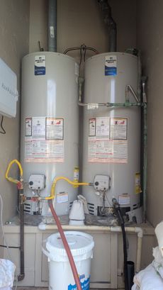 Two water heaters in a utility space. Yellow gas lines, white bucket, and pipes are visible.