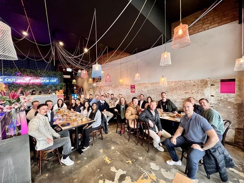 Large group of people gathered indoors at a restaurant. Some are seated, others are smiling. Dimly lit, colorful decor.