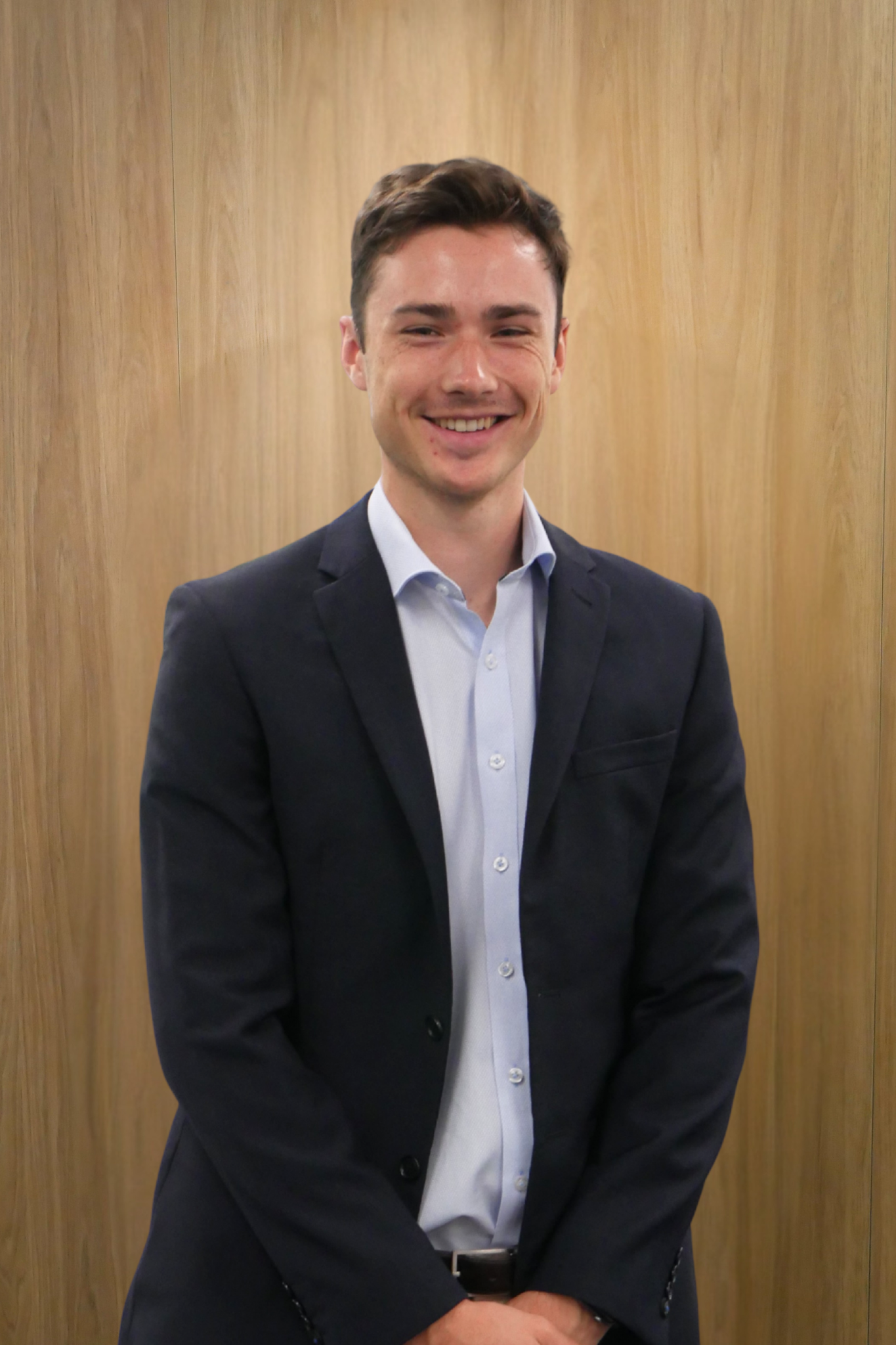 Man with a beard and a smile, wearing a navy blazer and red shirt, arms crossed, standing in front of a wood-paneled wall.