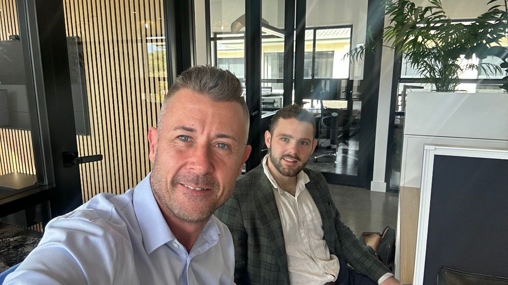 Two men smile at the camera in an office setting. One wears a blue shirt; the other, a sweater.