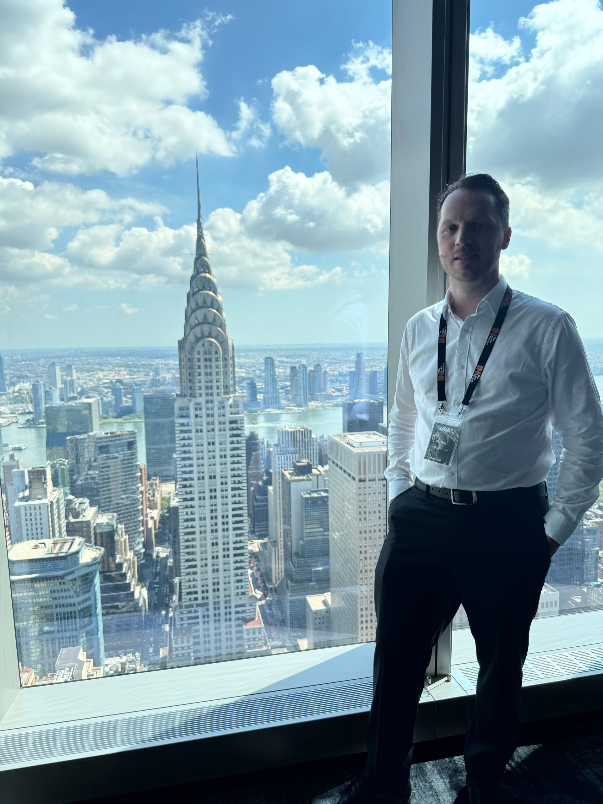 Man stands by window with the Chrysler Building in the background, overlooking a cityscape on a sunny day.