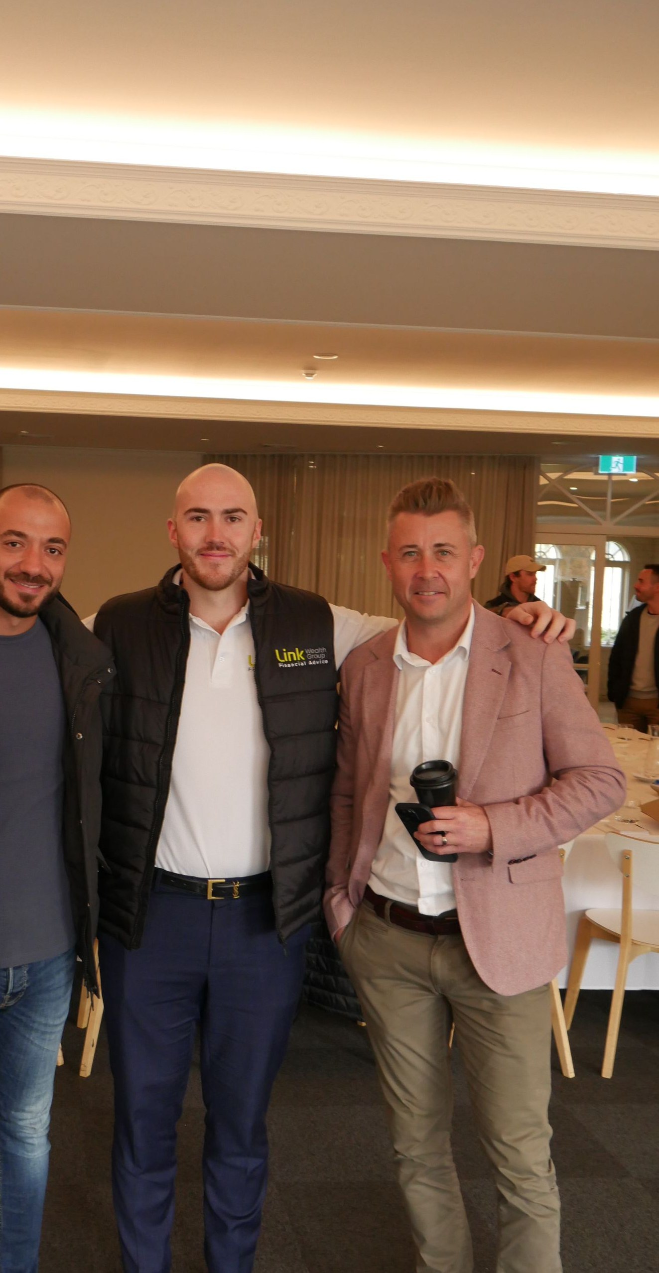 Three men pose indoors. One in a black vest, white shirt, and blue pants stands with a man in a pink blazer.