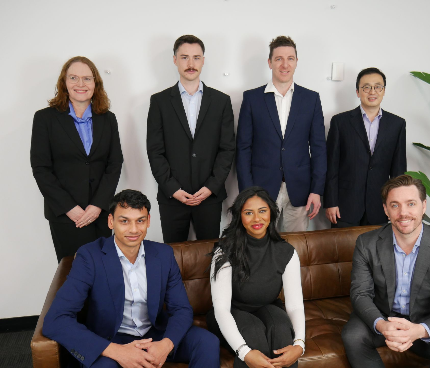 Group of eight professionals posing for a photo. Some are standing, others seated on a brown couch.