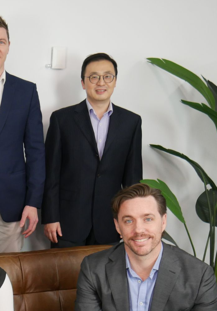 Three business people posing. A man in glasses in the center. Two in suits smiling. A plant is behind them.